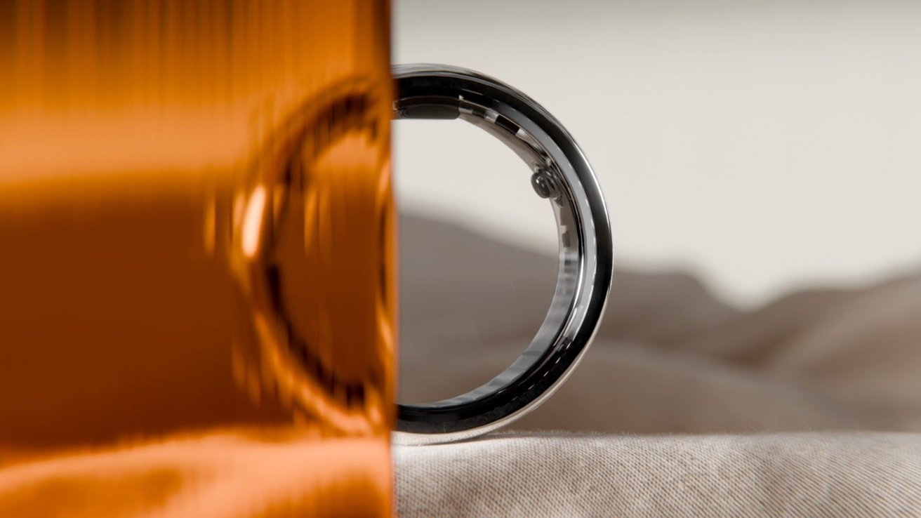 A silver ring partially obscured by an orange reflective surface, with a blurred neutral background.