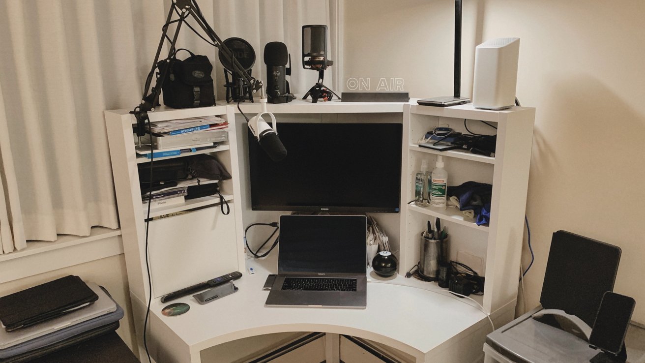 Workspace with white desk, laptop, monitors, microphones, shelves with books, sanitizer bottles, office supplies, and an 'ON AIR' sign.