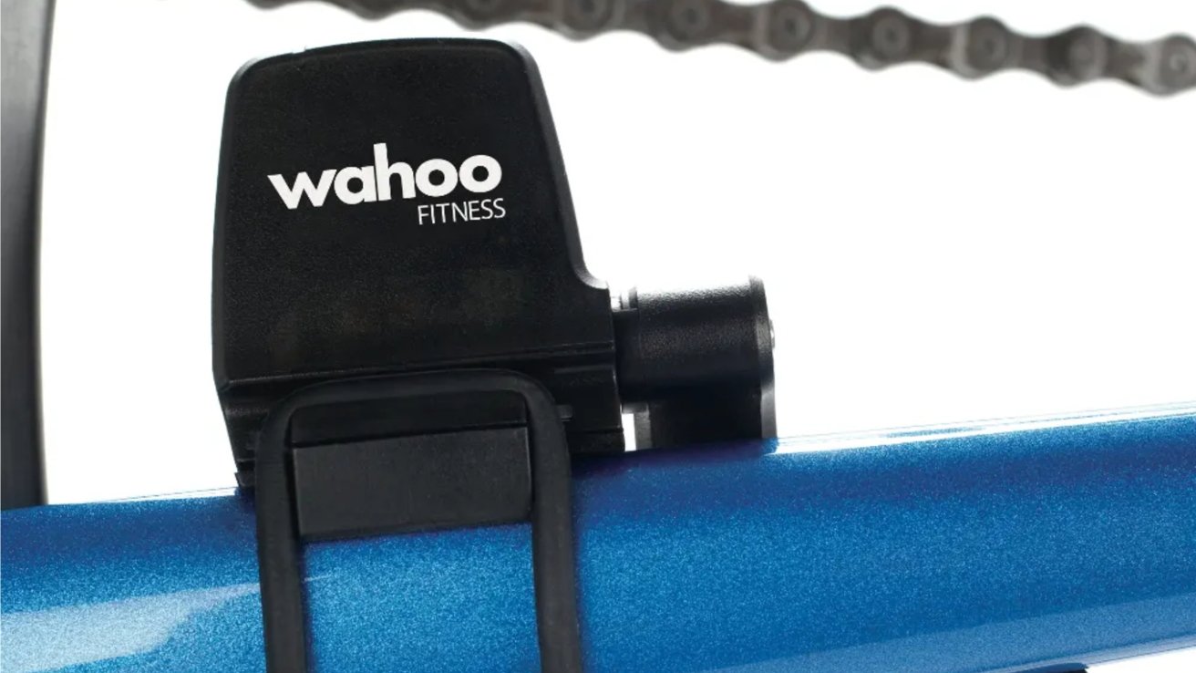 Perangkat Black Wahoo Fitness menempel pada permukaan biru metalik dengan rantai sepeda kabur di latar belakang.