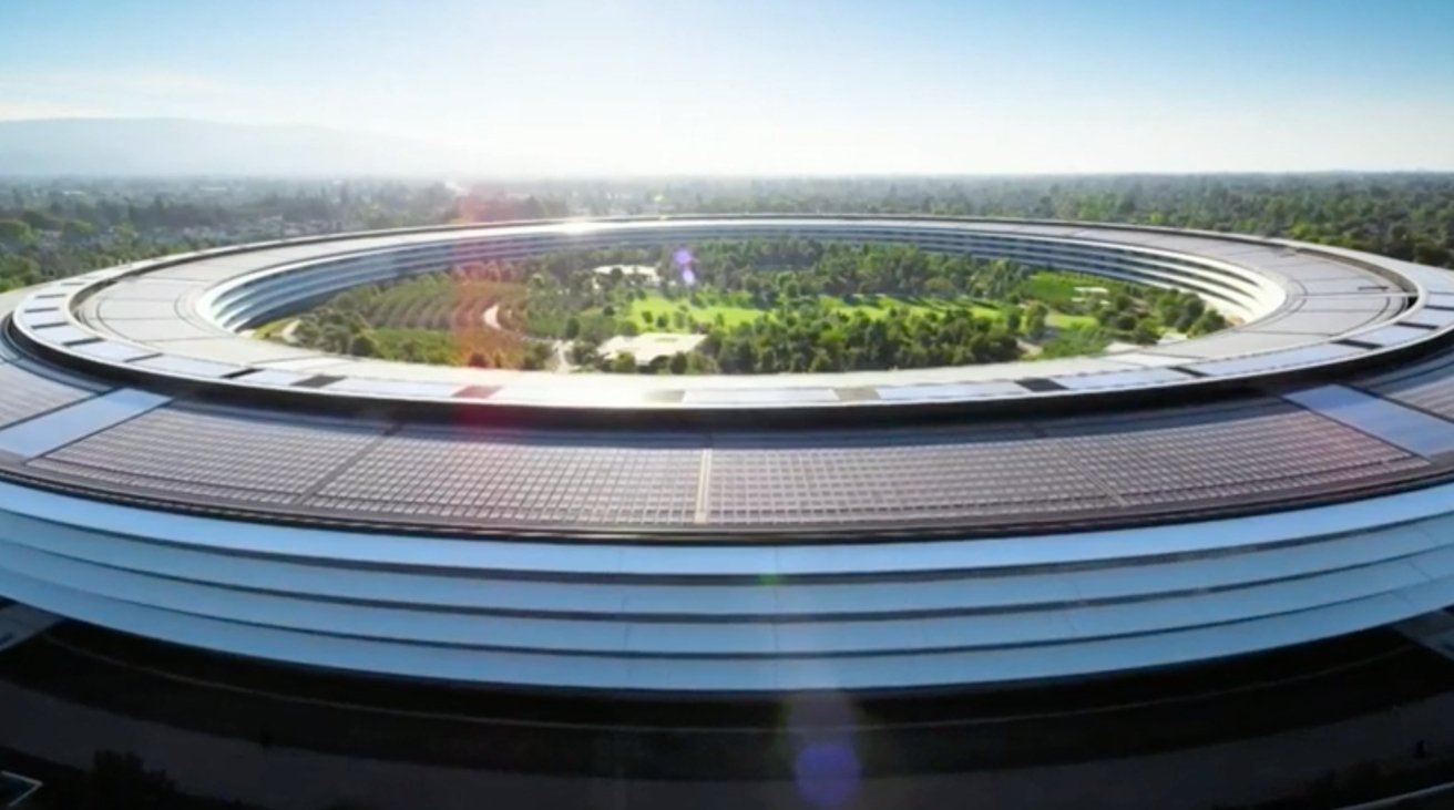 Circular building with a vast open center filled with green trees and pathways, surrounded by sleek glass and metal structure, under a clear blue sky.