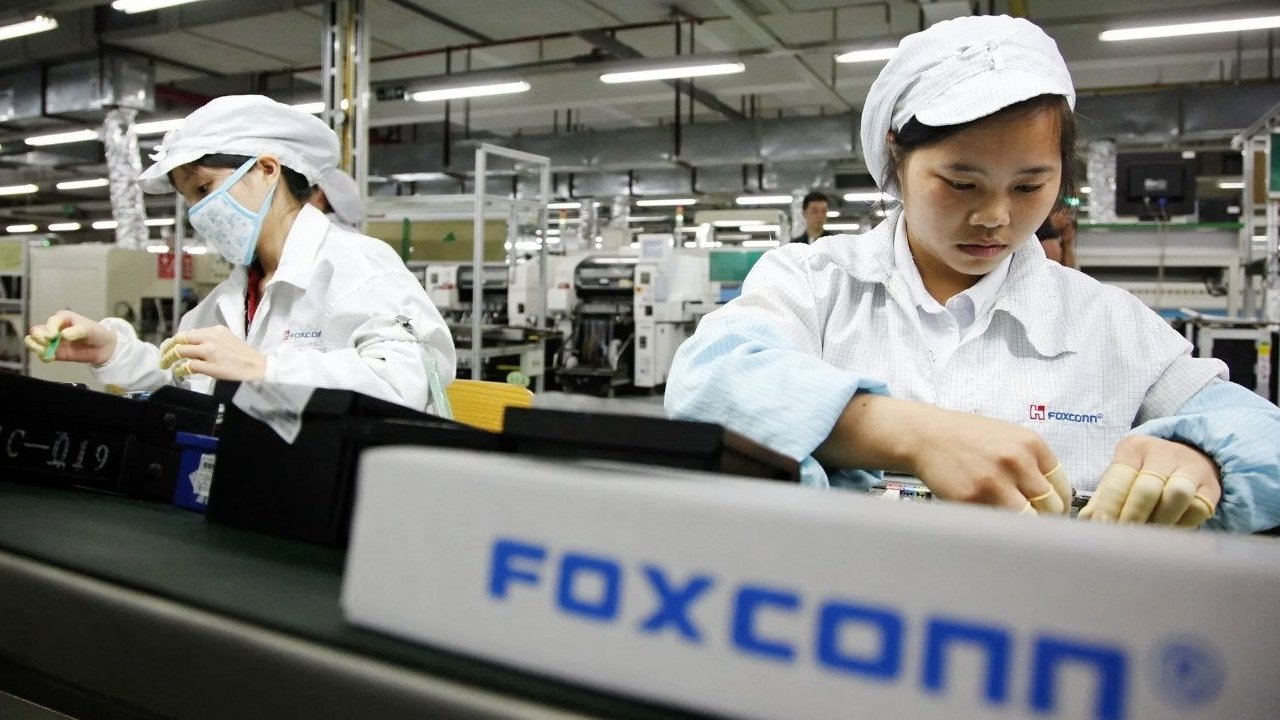 Two factory workers wearing white uniforms and caps assemble electronics on a production line. One worker wears a face mask, and the environment appears industrial and busy.