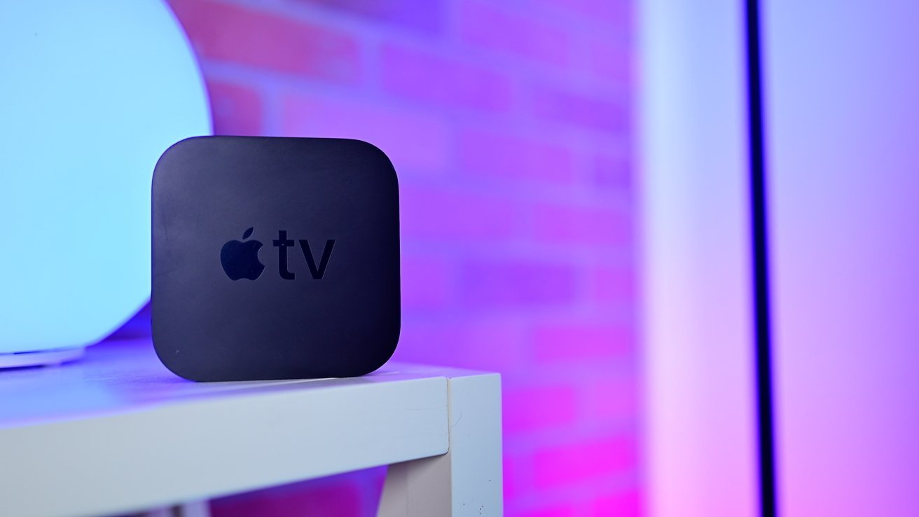 Black Apple TV device on a white shelf, with a colorful, out-of-focus brick wall background illuminated in blue and pink hues.