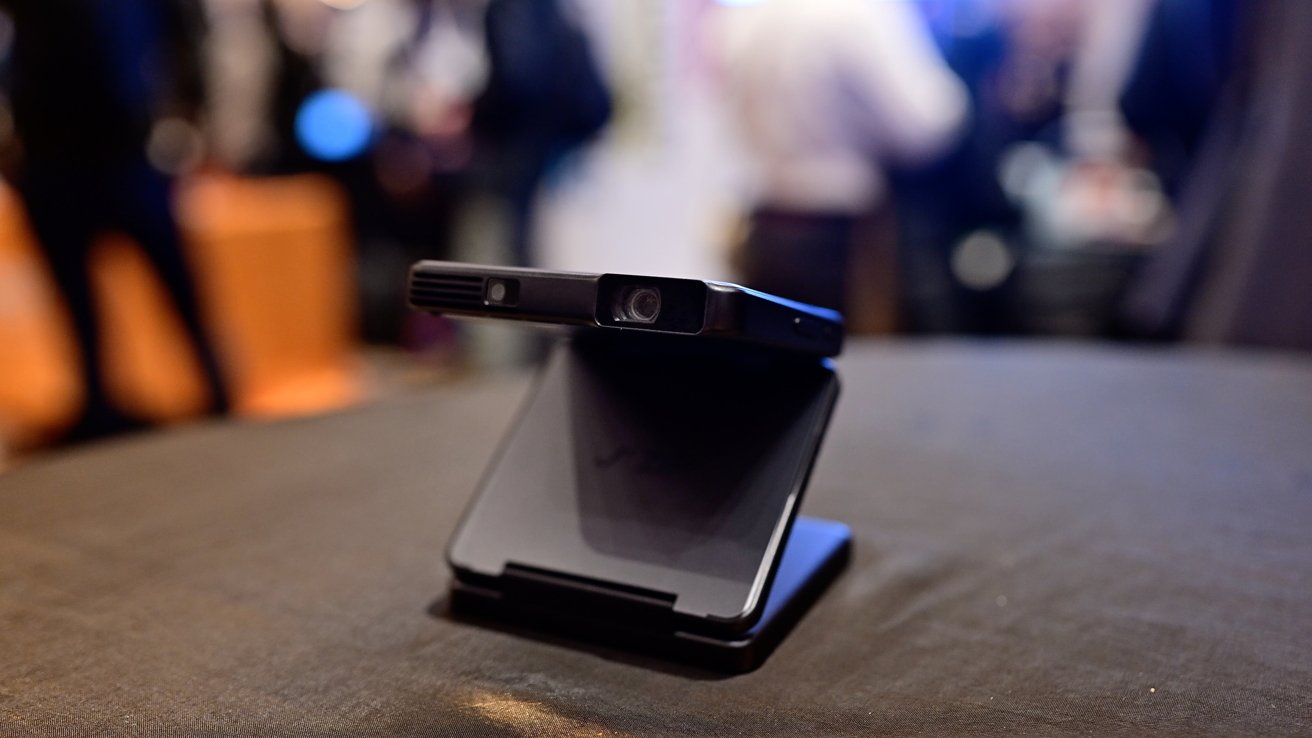 A black foldable smartphone with a hinge is displayed on a table, featuring a camera module on top, against a blurred background.