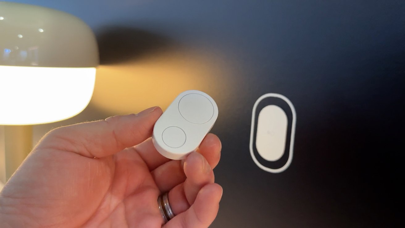 A hand holding a small white smart button next to its wall-mounted holder, with a glowing lamp nearby.