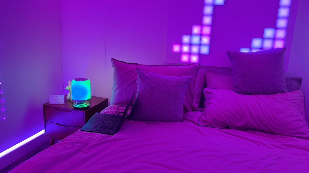 Bedroom with purple lighting, bed with pillows, colorful geometric wall lights, small bedside table with a cylindrical lamp, and an open laptop.