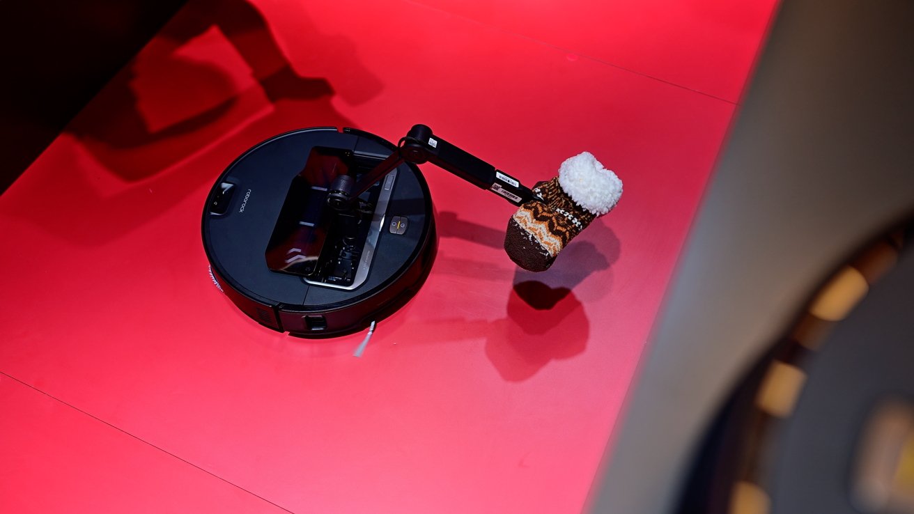 Robot vacuum with an extendable arm holding a patterned sock, placed on a vibrant red surface.