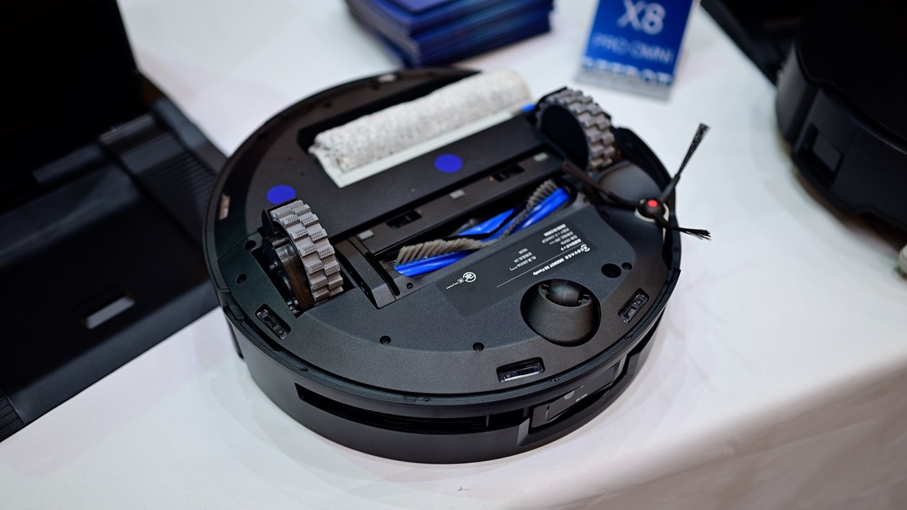 A round robotic vacuum cleaner on a table, showing its underside with wheels, brushes, and sensors.