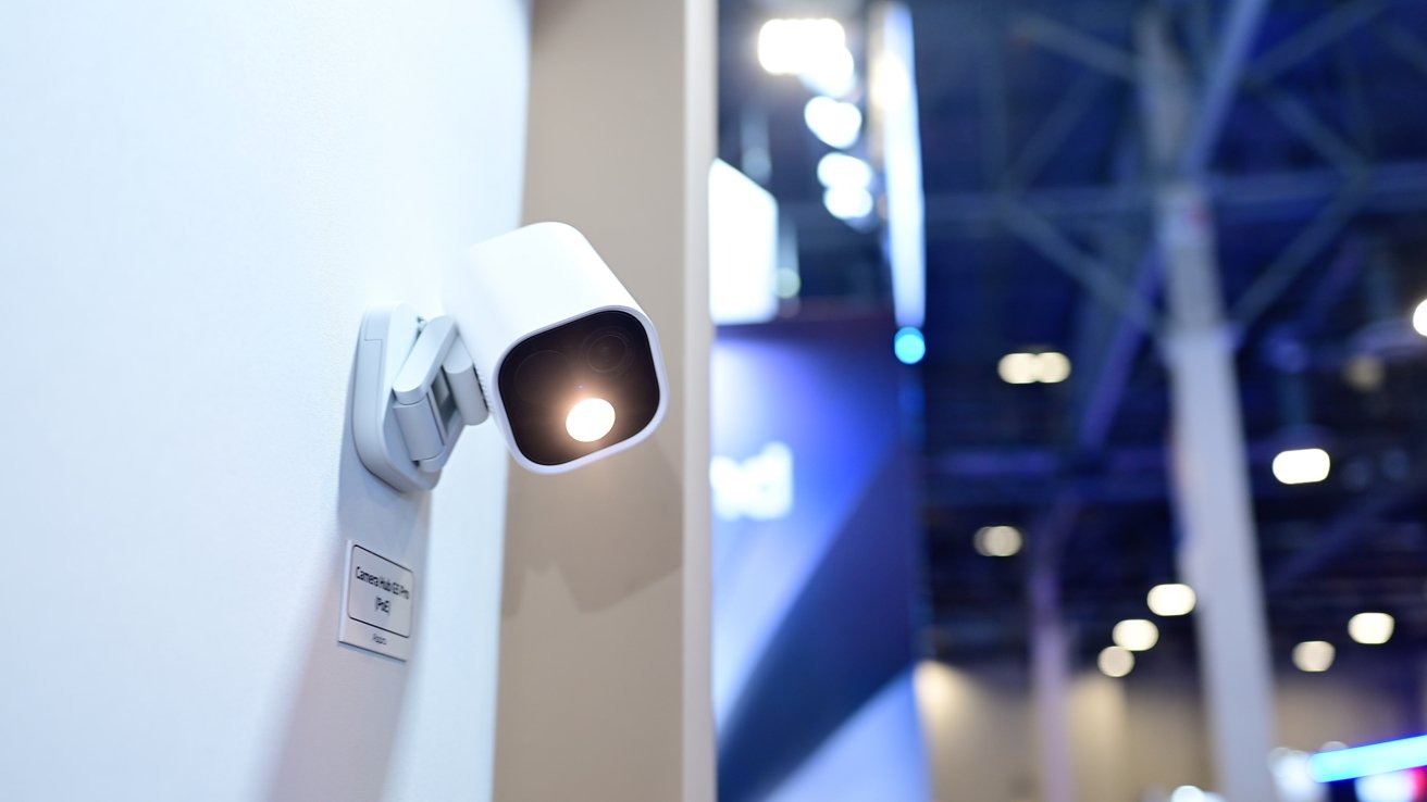 White security camera with a glowing light mounted on a wall, with blurry exhibition hall background featuring bright overhead lights and structural beams.