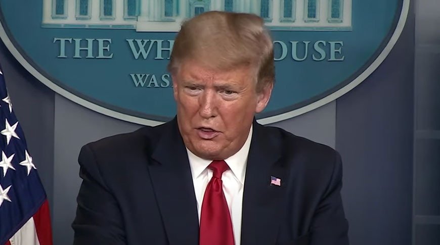 A man in a suit with a red tie speaks at a podium, flanked by an American flag and a sign reading 'The White House, Washington.'