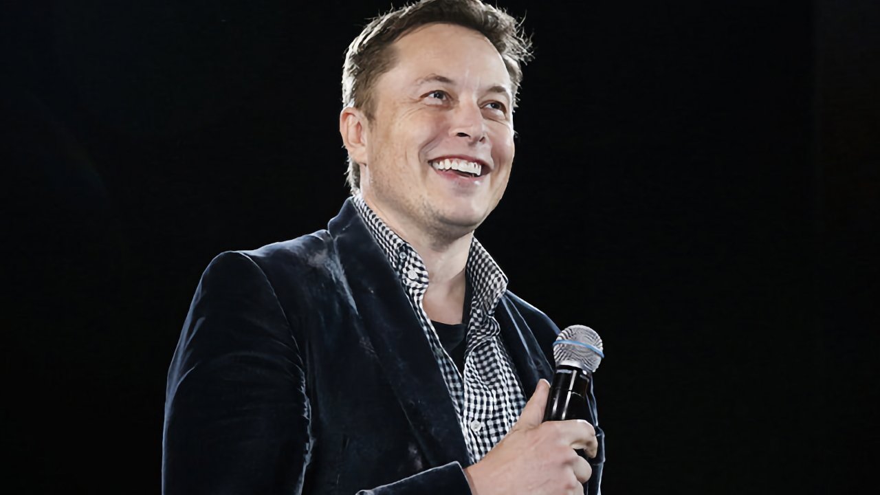 A person with short hair smiling while holding a microphone, wearing a dark jacket over a checkered shirt against a dark background.