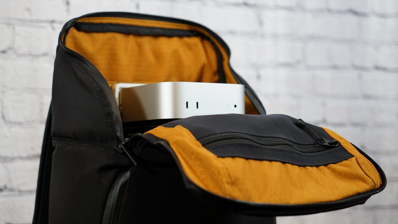 Open black backpack with orange interior containing a small silver computer against a white brick wall background.