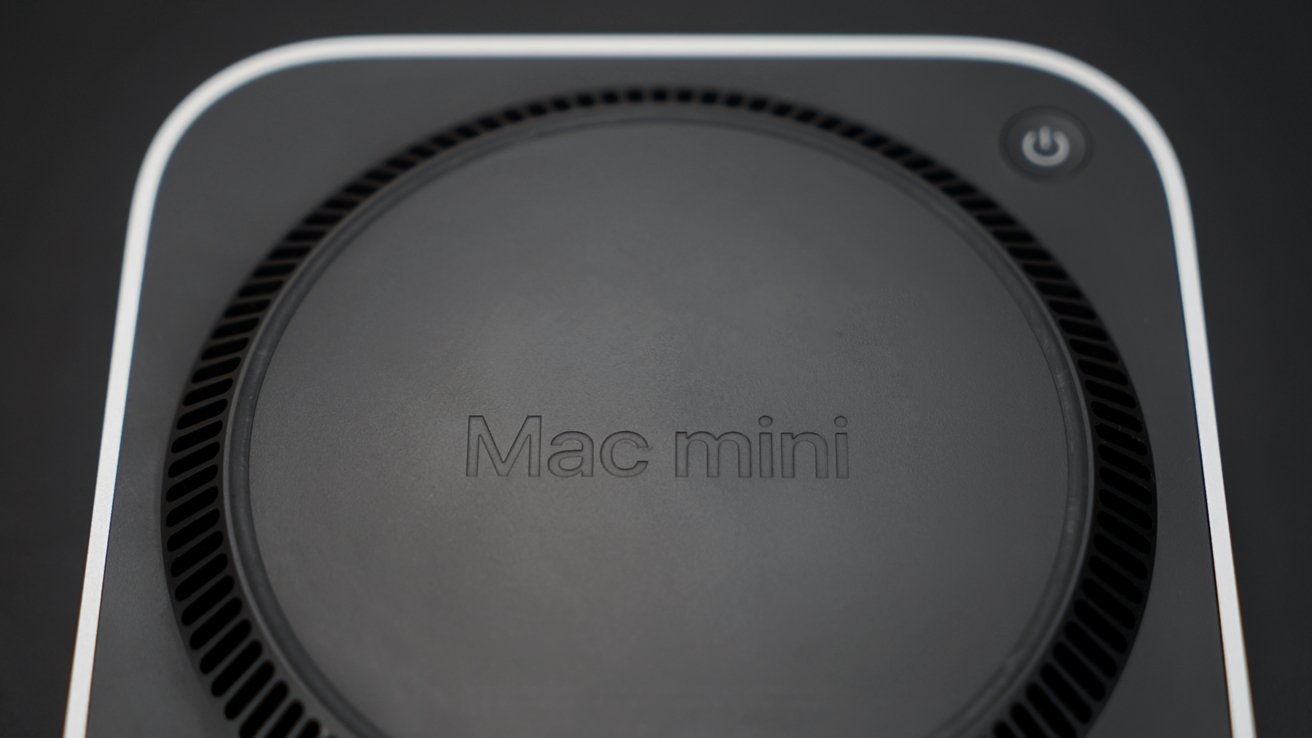 Close-up of a black circular Mac Mini with vented edges, featuring a power button icon in the upper right corner.