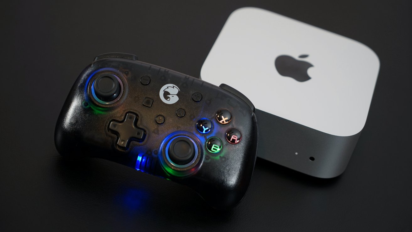 A black gaming controller with multicolored backlit buttons in front of a small, grey Apple device on a dark surface.