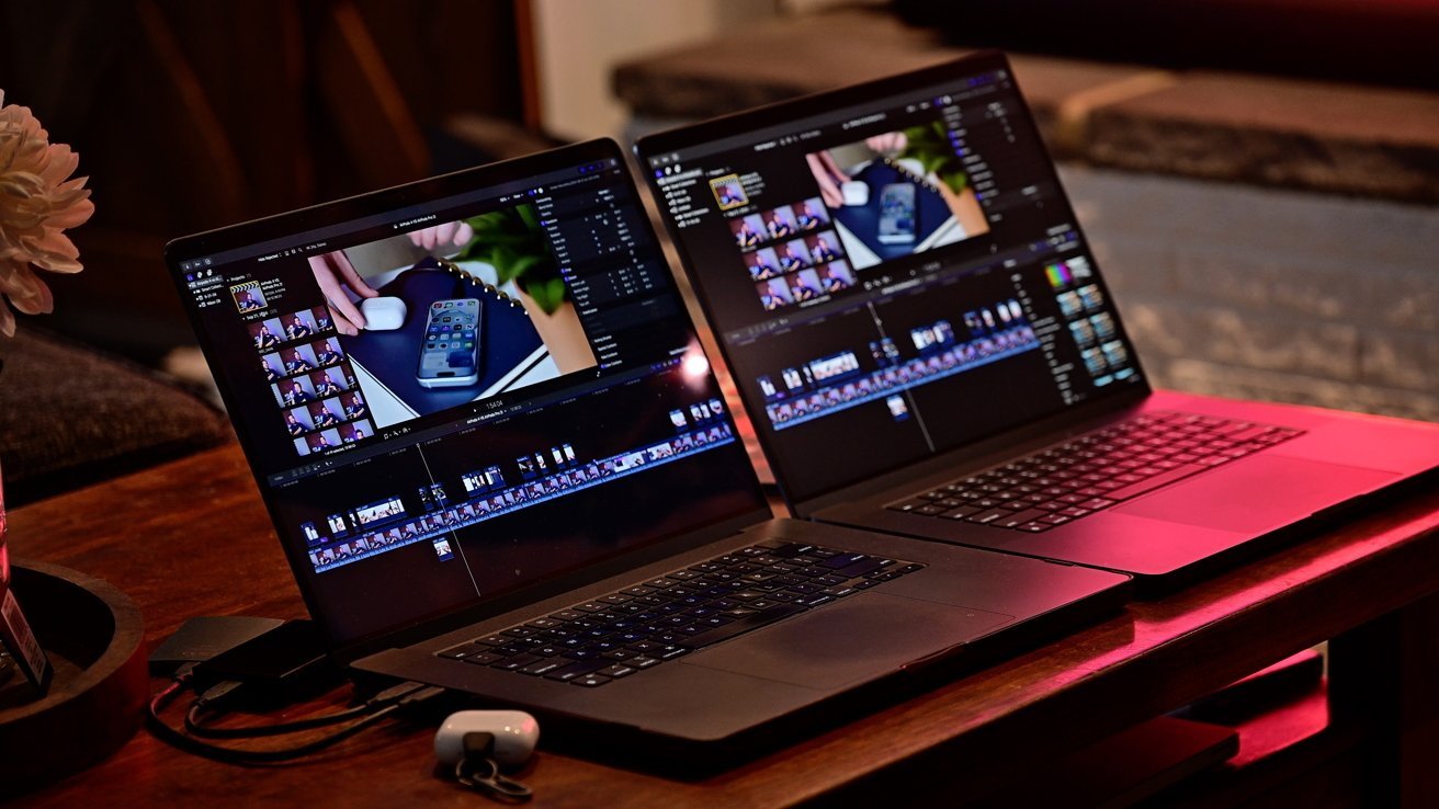 Two 16-inch MacBook Pro laptops on a wooden table displaying video editing software. A small case and a partial view of a white flower nearby. Warm lighting in the background.