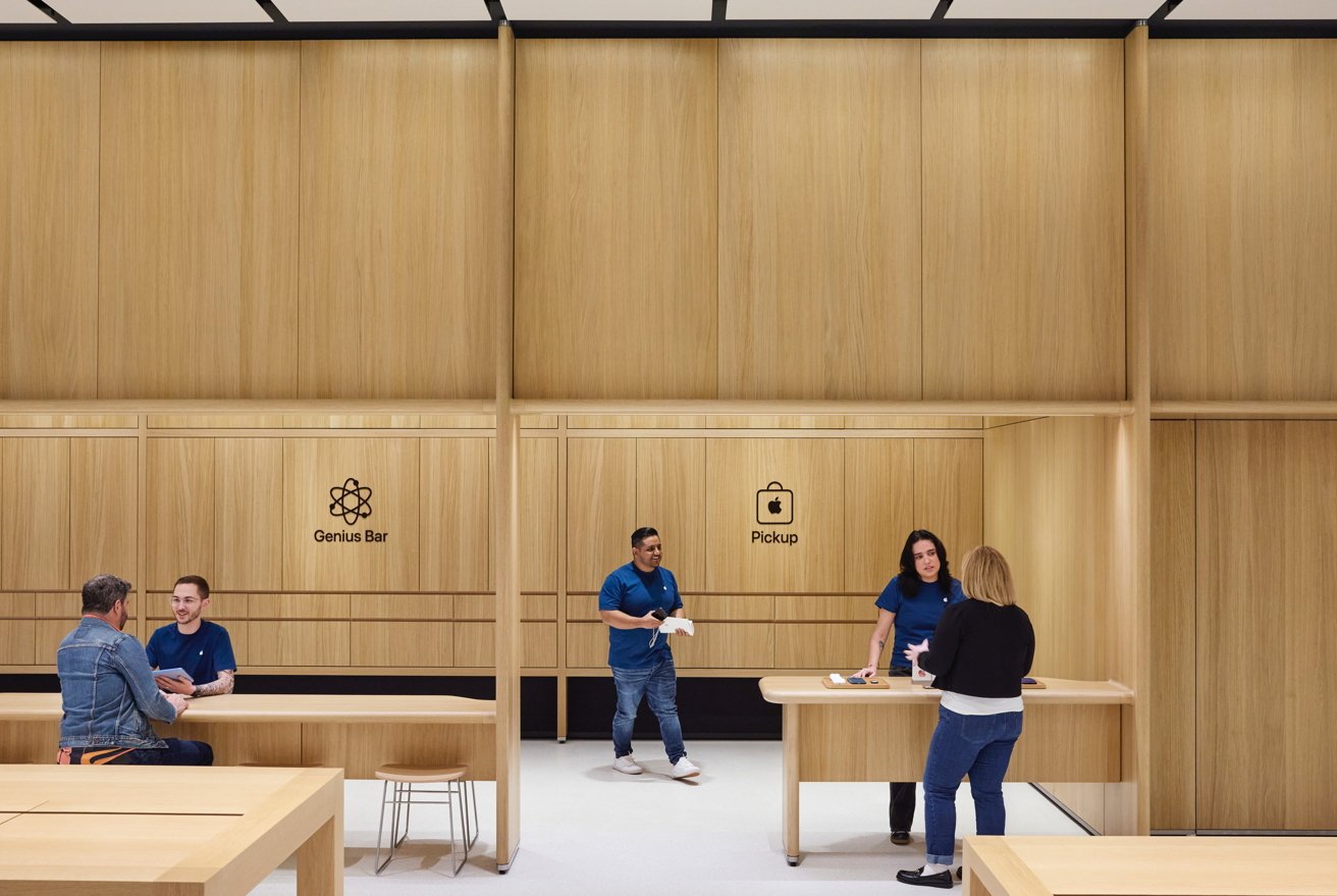 Store interior with light wood, two service counters labeled Genius Bar and Pickup, four people interacting, two seated, two standing.