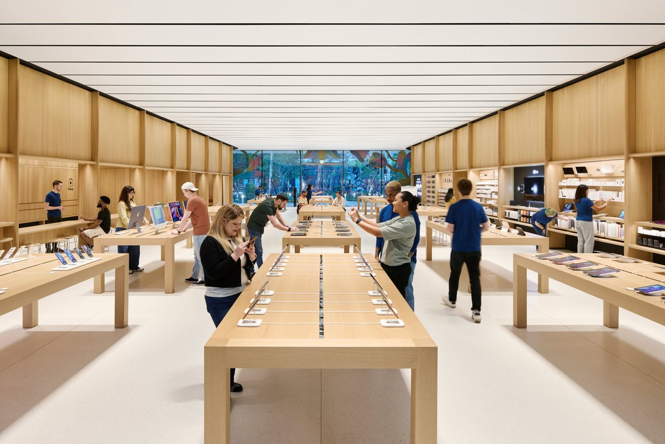 A spacious, bright tech store with wooden tables displaying electronic devices, several people browsing products, and staff in blue shirts assisting customers.