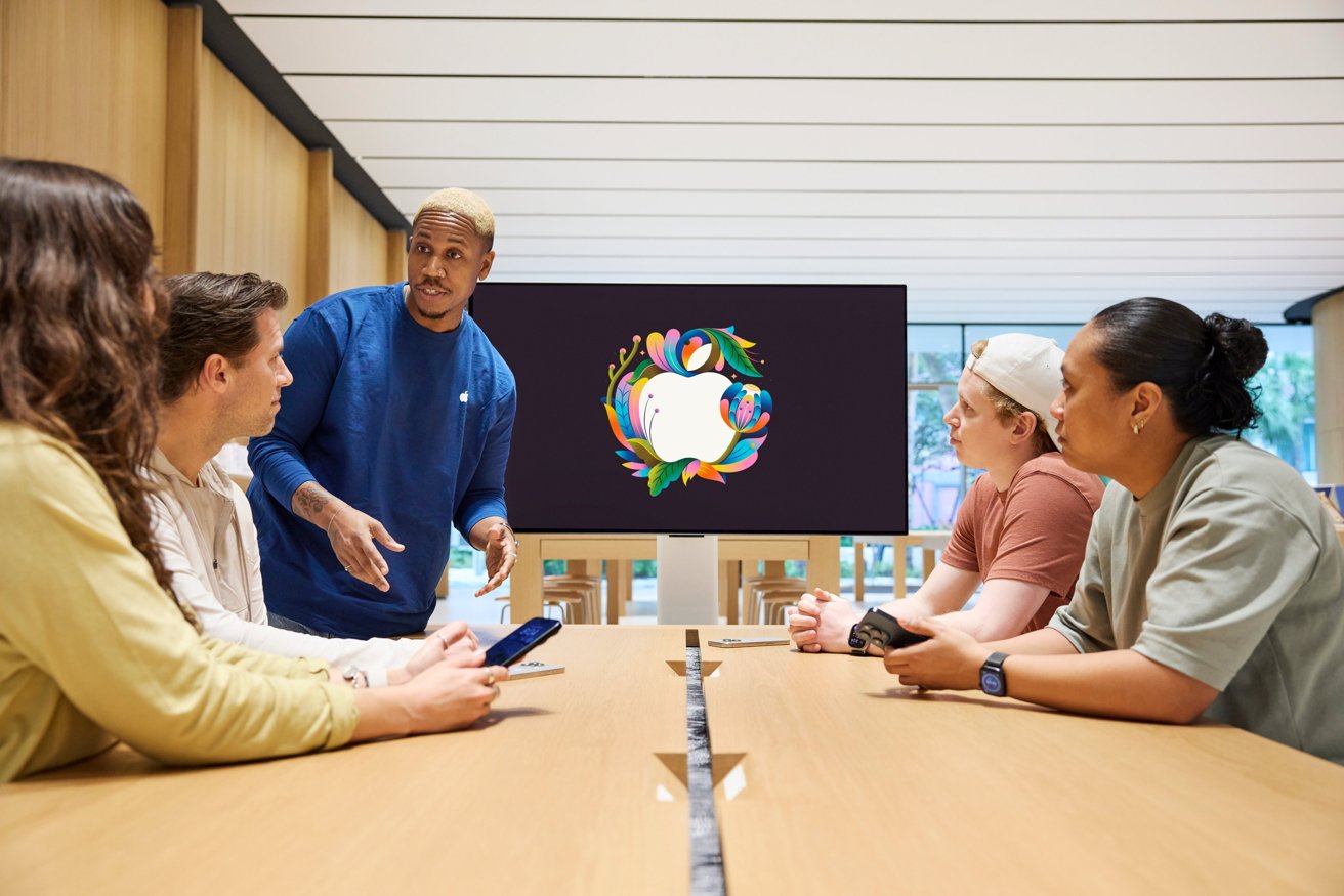 A group of five people sit at a table with a colorful Apple logo displayed on a screen behind them.