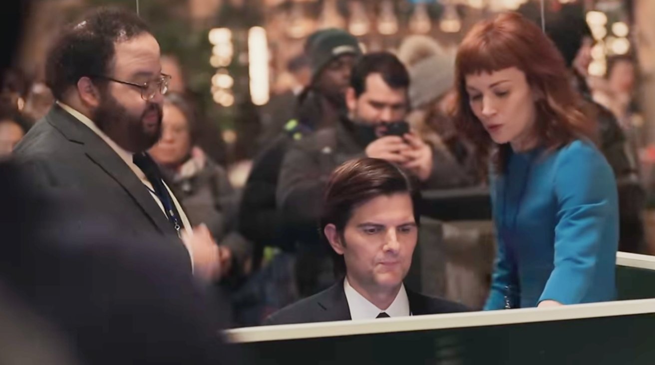 Man in a suit sits at a desk, woman in blue stands beside him, bearded man standing nearby, several people in background observing.