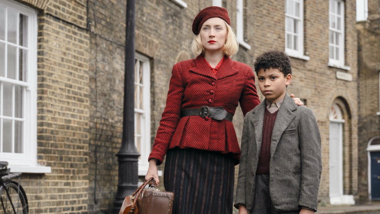 A woman in a red coat and beret stands beside a boy in a gray suit, outside brick buildings. She holds a brown suitcase.