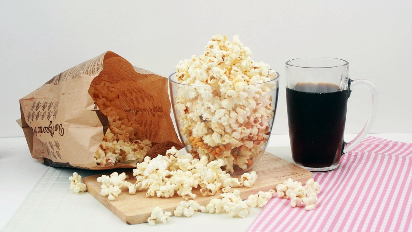 A paper bag and glass bowl filled with popcorn, next to a glass mug of dark beverage on a pink striped cloth.