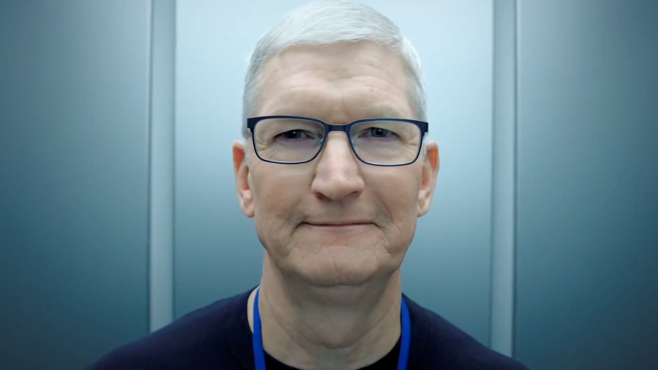 Smiling older man with short white hair and glasses, wearing a black shirt, standing against a plain, softly lit background.