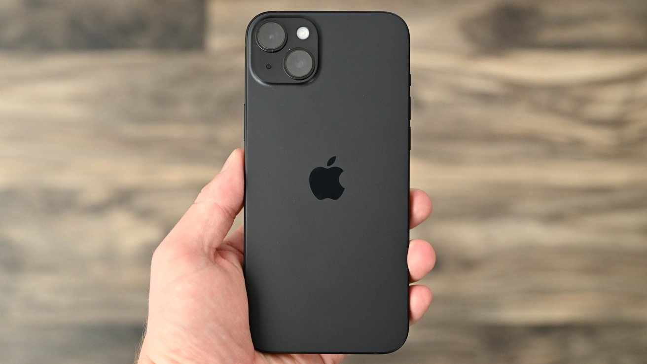 Hand holding a black iPhone 15 showing the back with dual rear cameras and an Apple logo, on a blurred wooden floor background.