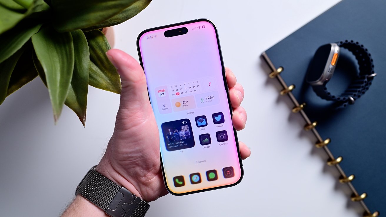 Person holding a smartphone with calendar and app icons, next to a notebook, smartwatch, and plant on a white surface.