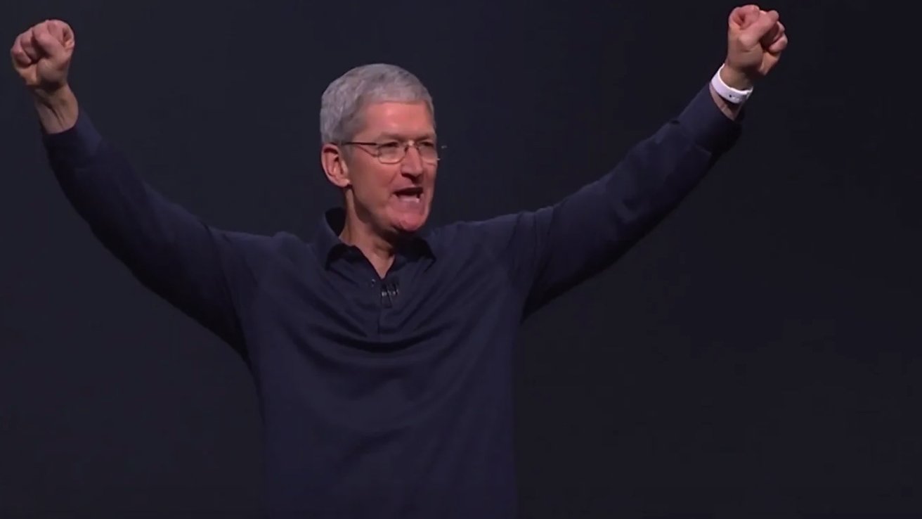 Tim Cook, with short gray hair and glasses raises both arms enthusiastically, wearing a dark shirt against a plain background.