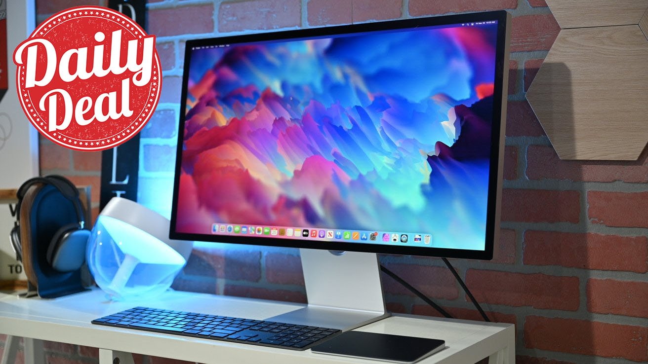 Desktop setup with Apple Studio Display, keyboard, and Magic trackpad, headphone stand, ambient lighting, red 'Daily Deal' label, and hexagonal wall decor in brick-walled room.