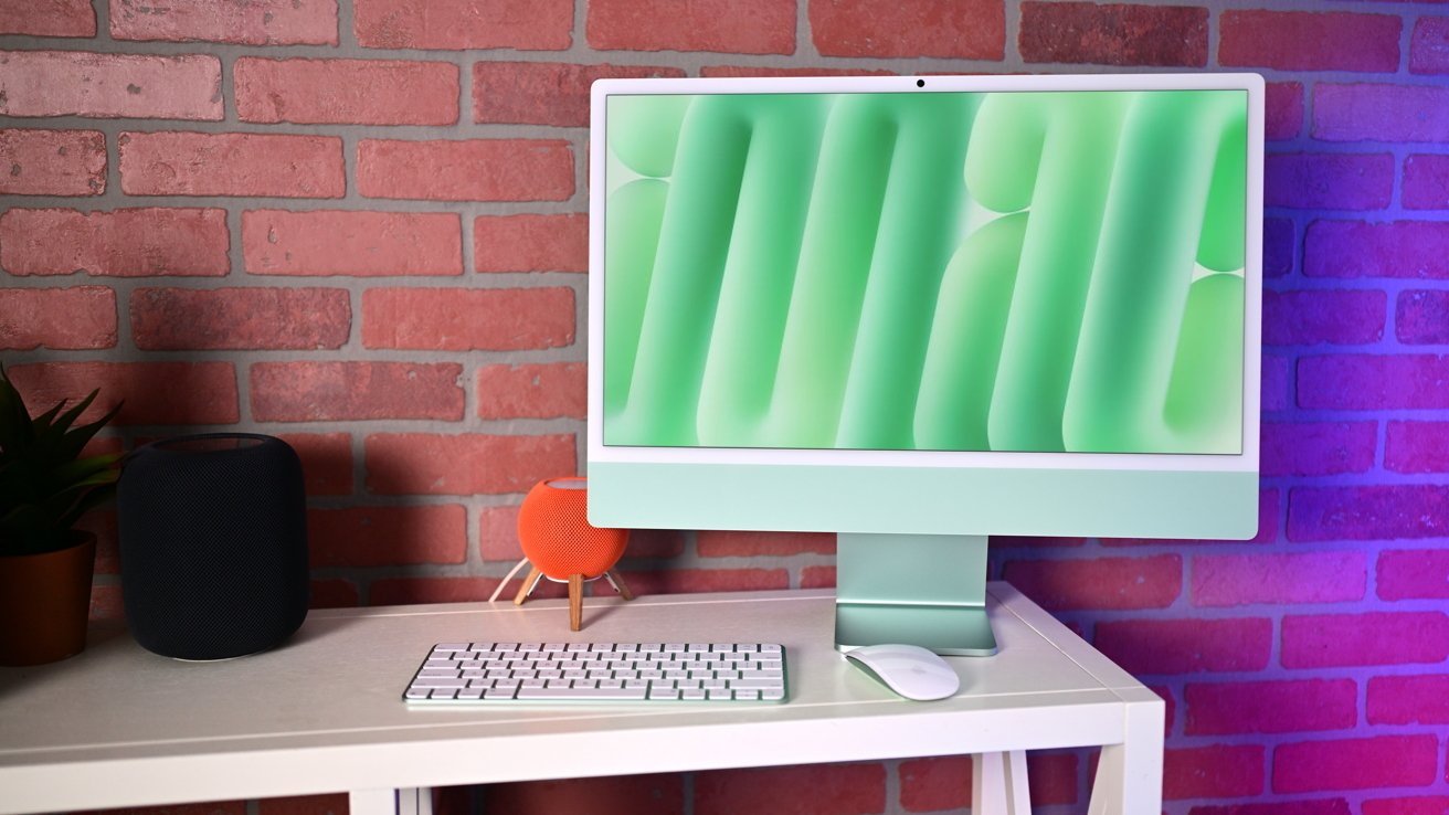 Apple 24-inch iMac M4 desktop computer with a green screen, keyboard, and mouse sits on a white table. Nearby are a black HomePod 2 speaker, orange HomePod mini speaker, and potted plant, against a brick wall.