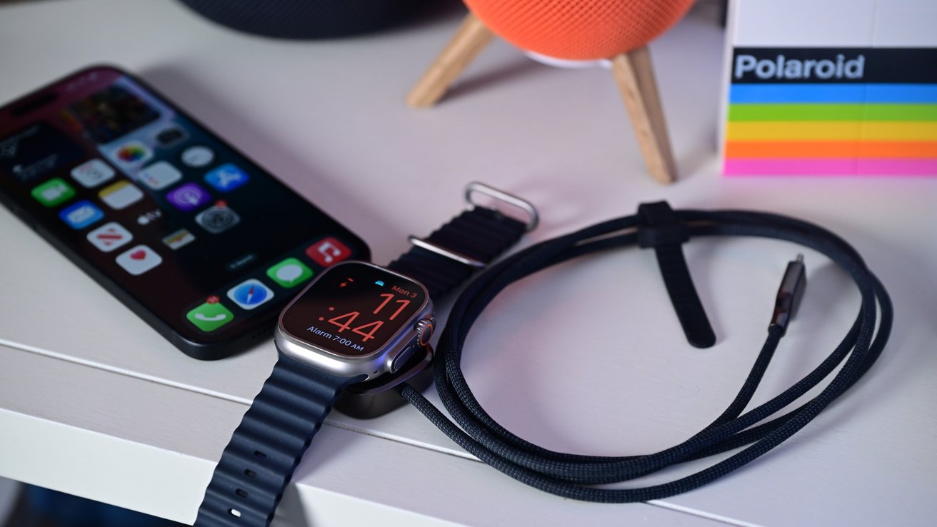 Smartphone with colorful apps, smartwatch displaying time, charging cable, orange speaker, and Polaroid-branded box on a white surface.