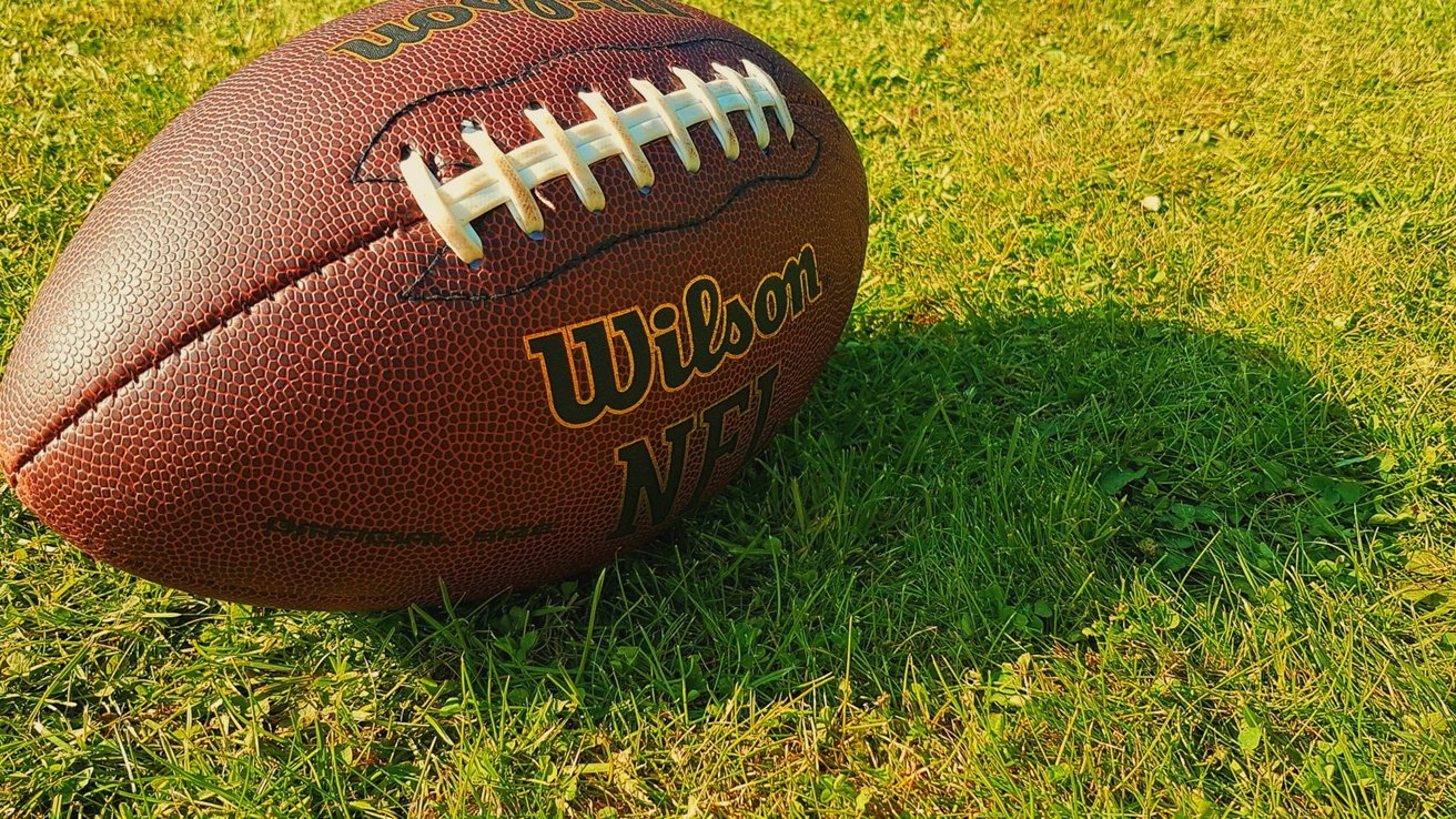 A brown American football rests on vibrant green grass, showing white laces and printed logo under bright sunlight.