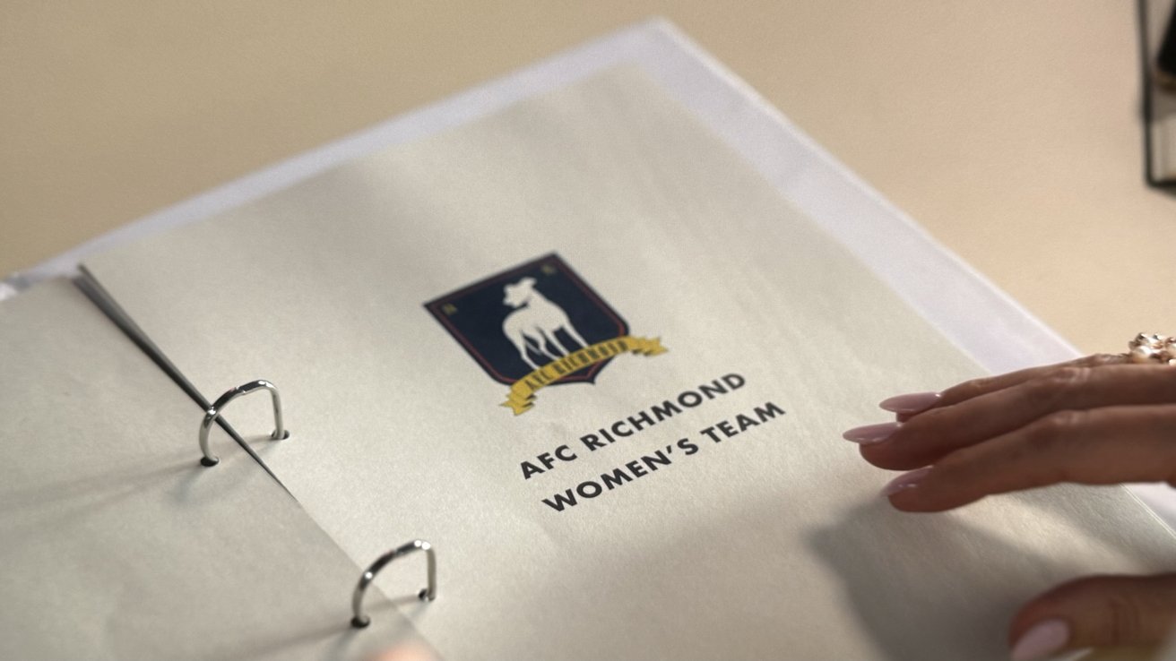 Binder with a cover reading AFC Richmond Women's Team, featuring a logo with a stylized greyhound. A person's hand with manicured nails is touching the page.