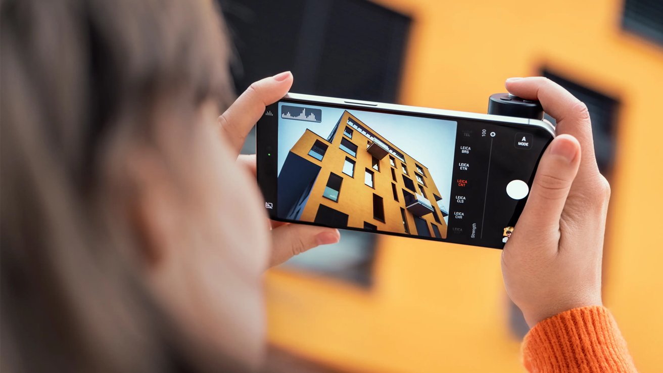 Person holding an iPhone with grip, capturing a modern, yellow building with angular windows, against a blurred background.