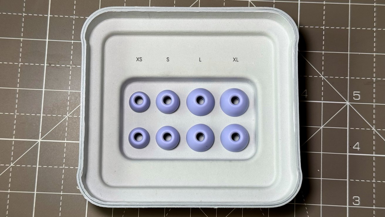 Packaging with eight lavender earbud tips in two rows, labeled XS, S, L, XL, arranged on a gray gridded mat.