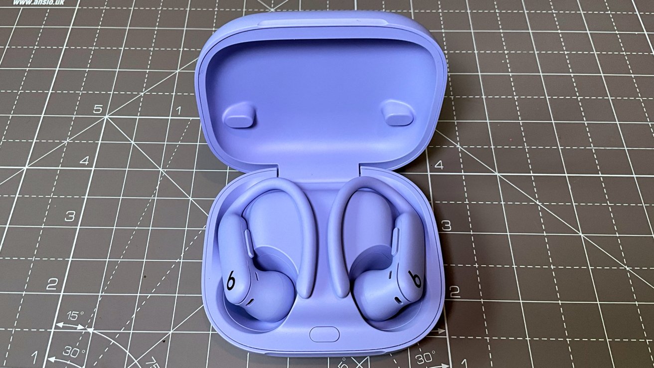 Purple wireless earbuds with ear hooks in an open, matching case resting on a grid-patterned surface.