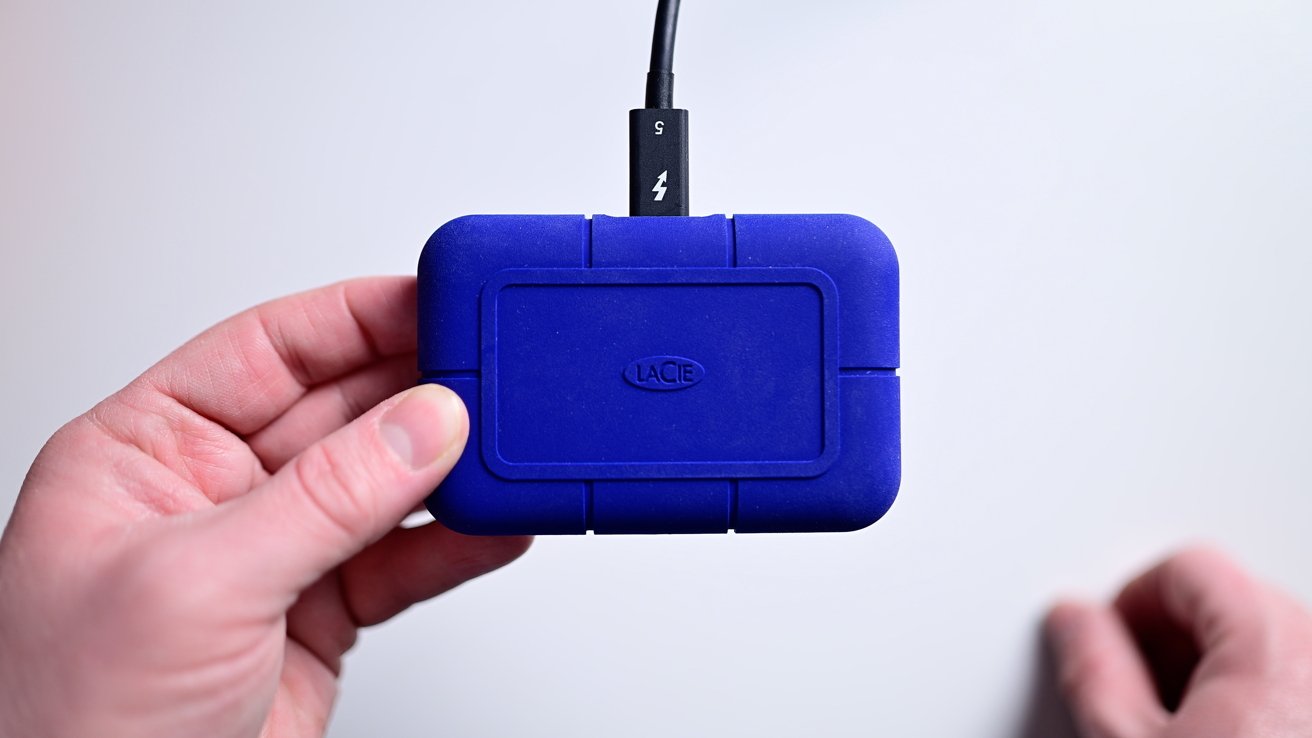 A‍ person holding a compact external hard drive with a⁤ cable against a neutral background.