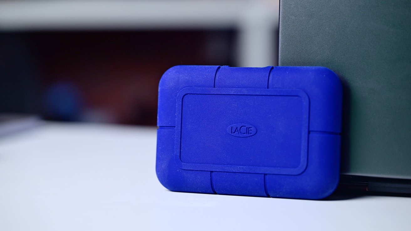 Blue rectangular hard drive with rounded edges sits on a white surface, partially leaning against a dark green object in the background.