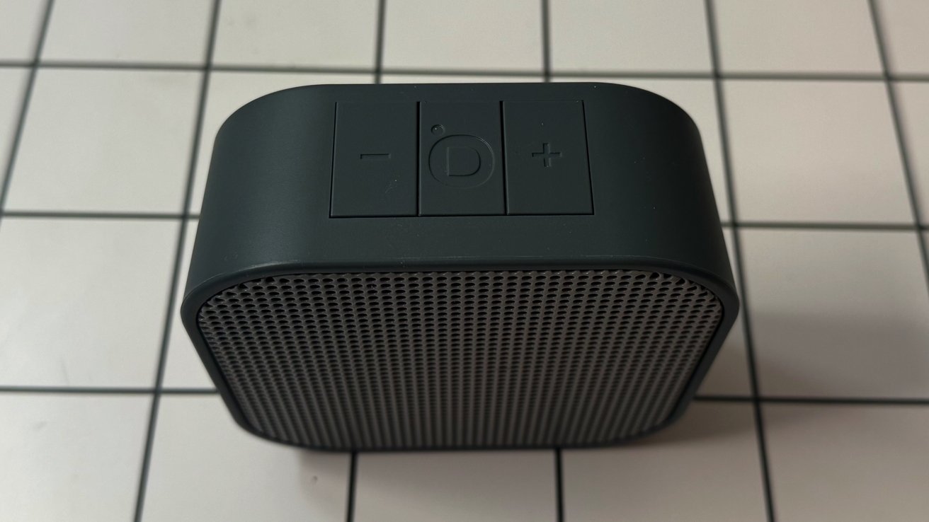 Speaker hitam kecil dengan gril pola grid, ditempatkan di permukaan ubin. Tombol teratas termasuk minus, daya tengah, dan simbol plus.