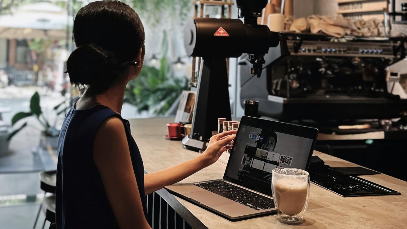 A person with hair in a bun uses a laptop in a cafe setting, next to an espresso machine and a glass of coffee.