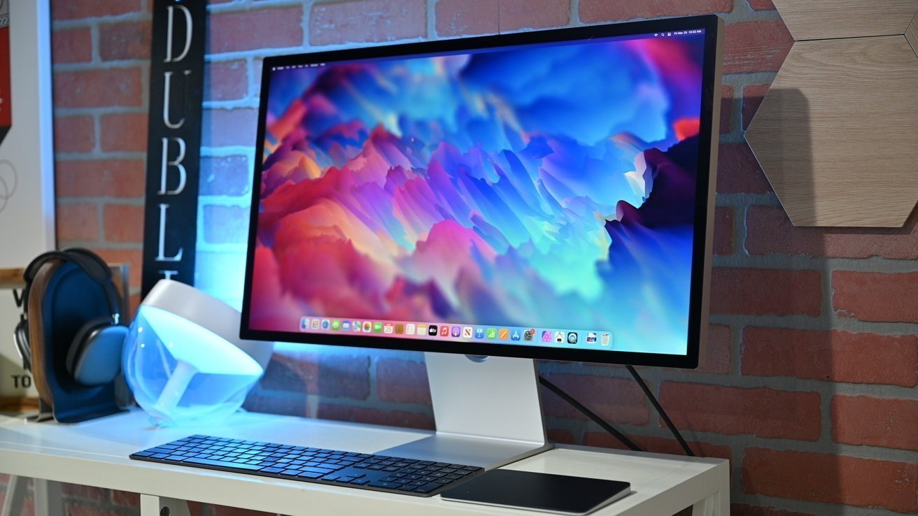 Computer monitor displaying colorful abstract art on a white desk with a keyboard, headphones, lamp, against a red brick wall background.