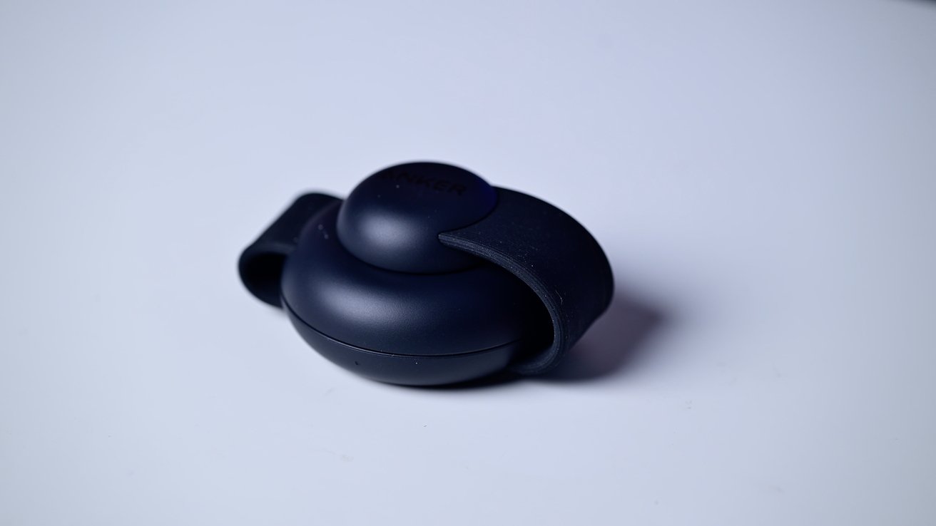 Circular black device with rounded top, resting on a white surface. The top has a raised dome with a looped strap.