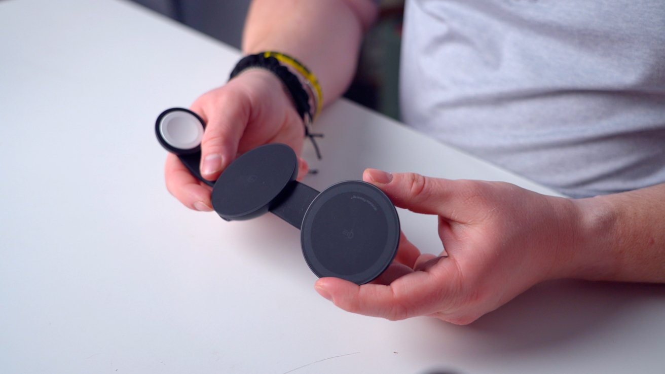 Hands holding a foldable wireless charging device with three black circular pads on a white table.