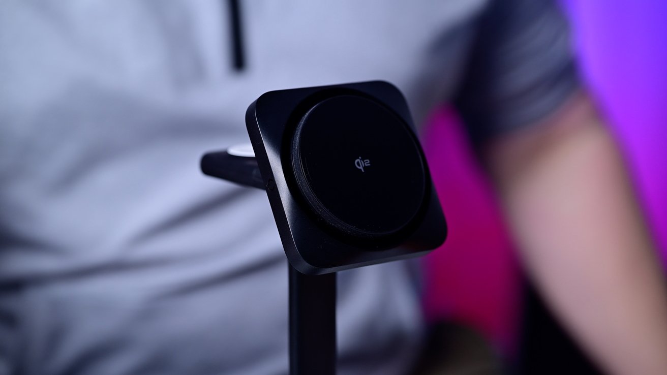 Close-up of a black wireless charging pad on a stand, with a blurred background featuring shades of purple and a person wearing a gray shirt.