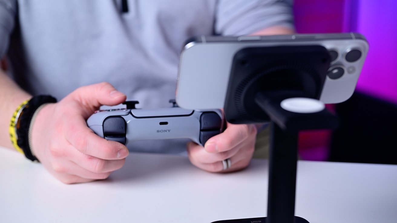 Person holding a grey PlayStation controller with a smartphone mounted on a stand nearby.