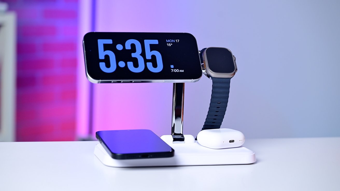 A multi-device charging station holds a smartphone, smartwatch, and wireless earbuds against a blurred purple and blue background.