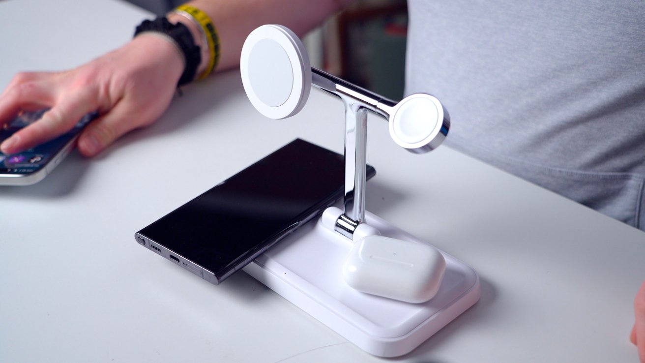 A wireless charging station with smartphone and earbuds on a white surface, near a person's hand holding another phone.