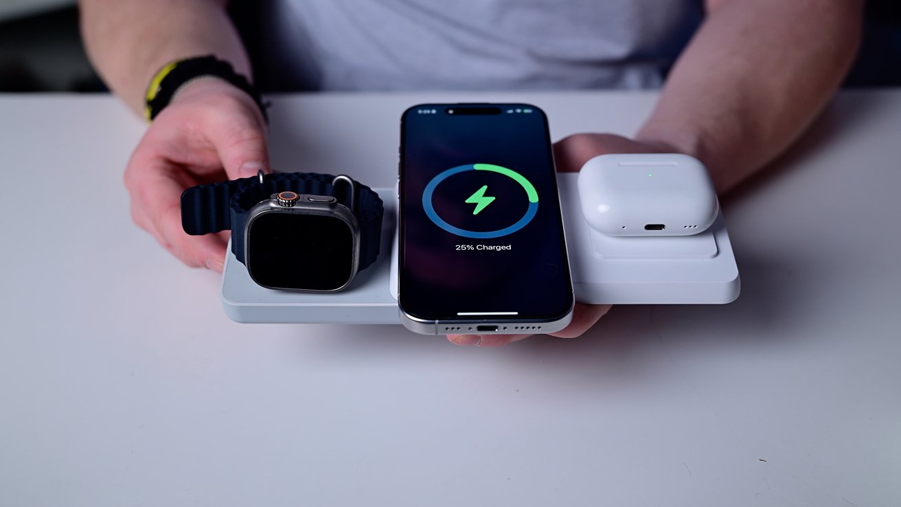 Smartwatch, smartphone, and wireless earphones charging on a white charging pad, with a charging status displayed on the smartphone screen, held by someone's hands.