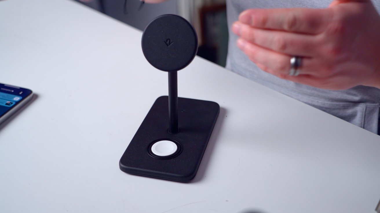 A person demonstrating a black wireless charging stand with a circular magnetic holder and smartphone slot on a table.