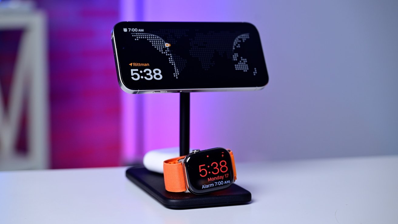 Smartphone and smartwatch on a black stand display digital clocks; orange watch band, blurred purple background.